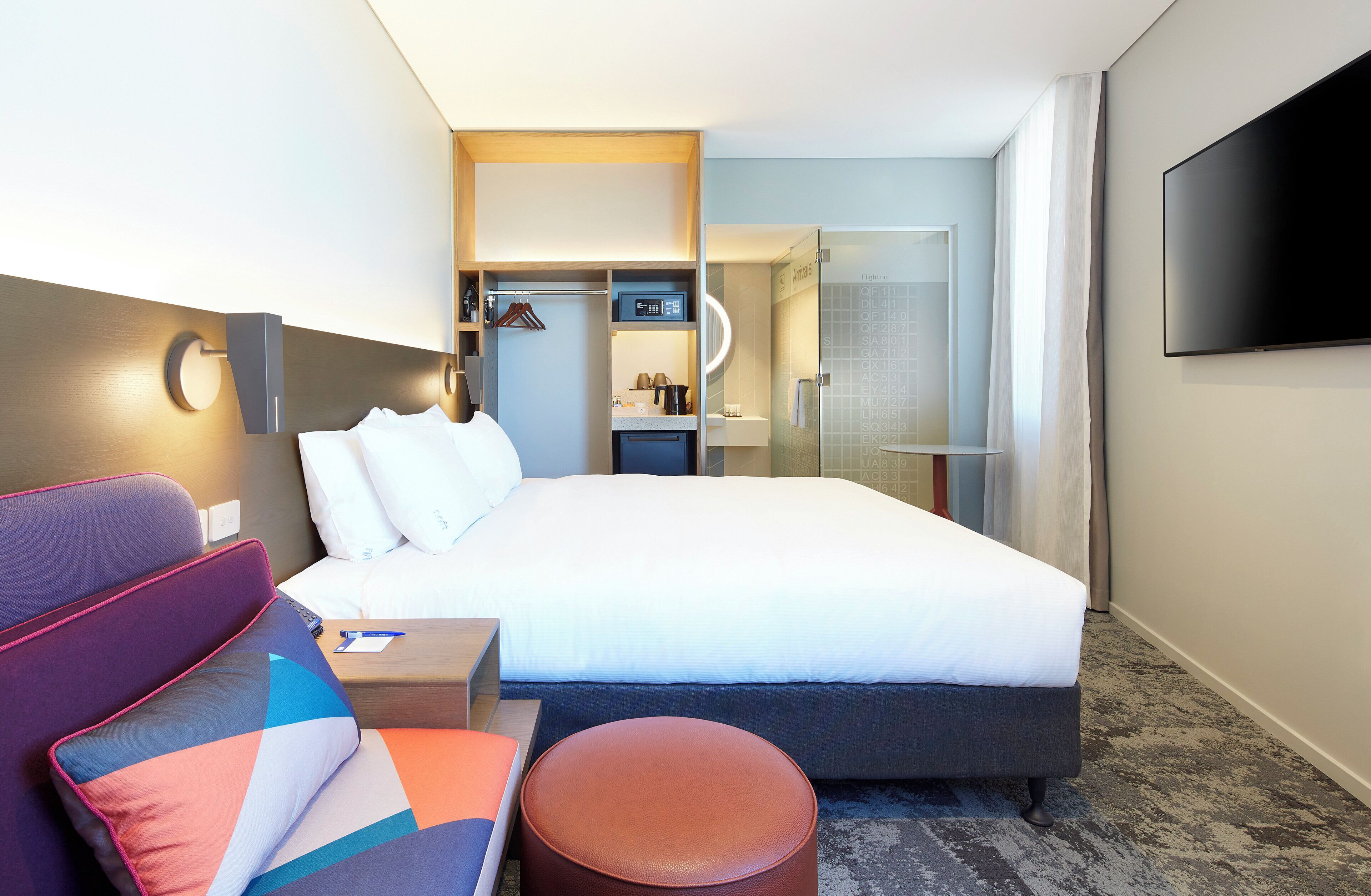 Une chambre d’hôtel moderne équipée d’un lit, d’un coin salon avec un canapé et un pouf, d’un téléviseur fixé au mur et d’une douche en verre.