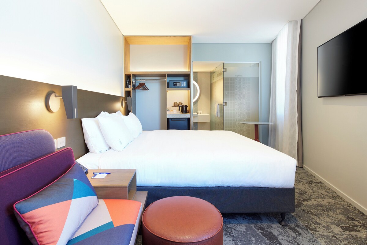 Een moderne hotelkamer met een bed, een zithoek met een bank en voetenbankje, een aan de muur bevestigde televisie en een douchecabine met glazen wanden.