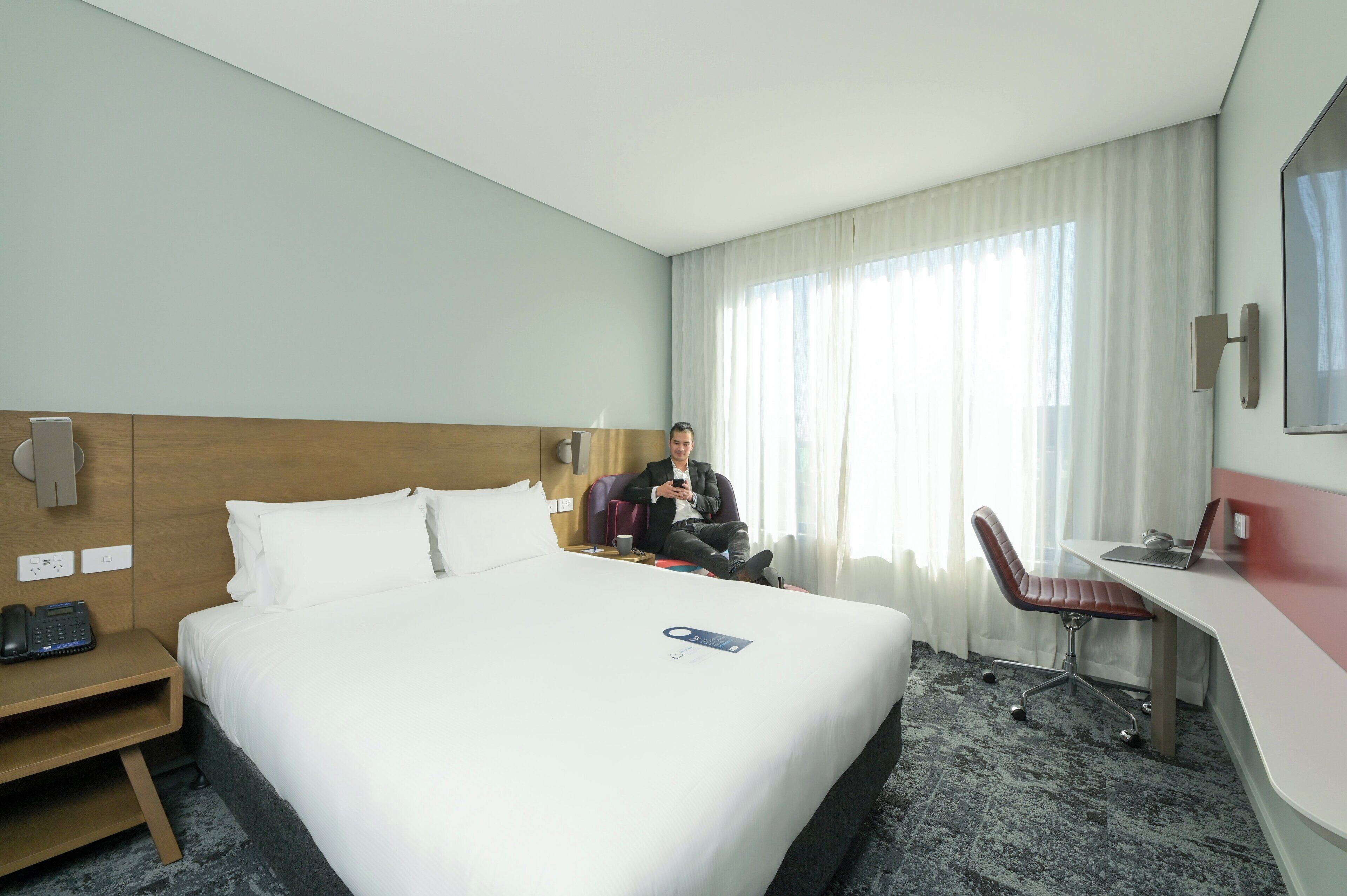 Una habitación de hotel con una cama grande, un escritorio con una computadora portátil, una silla, un teléfono en la mesita de noche y una ventana con cortinas.