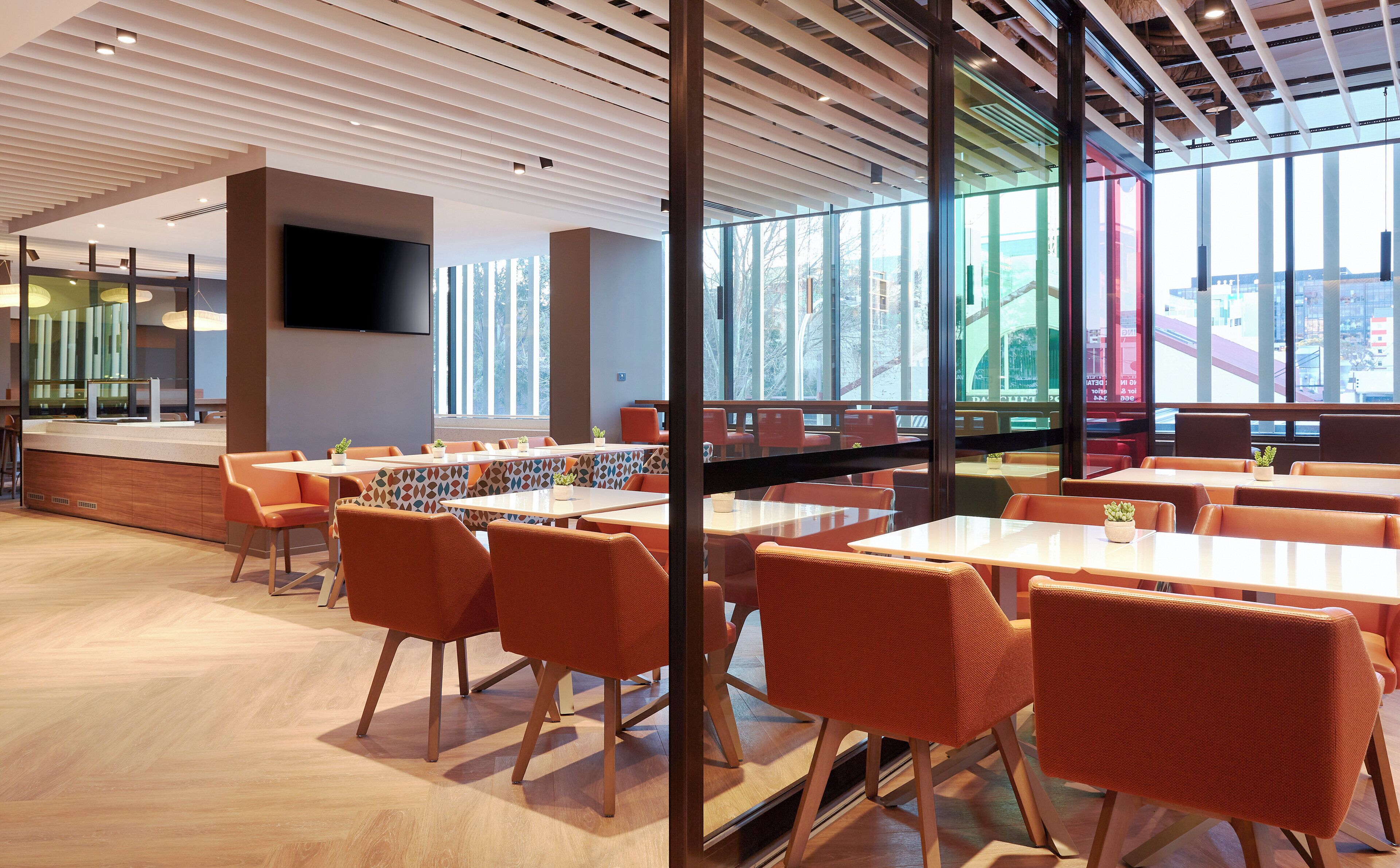 A modern dining area with tables and chairs, large windows, and a flat-screen TV mounted on the wall.
