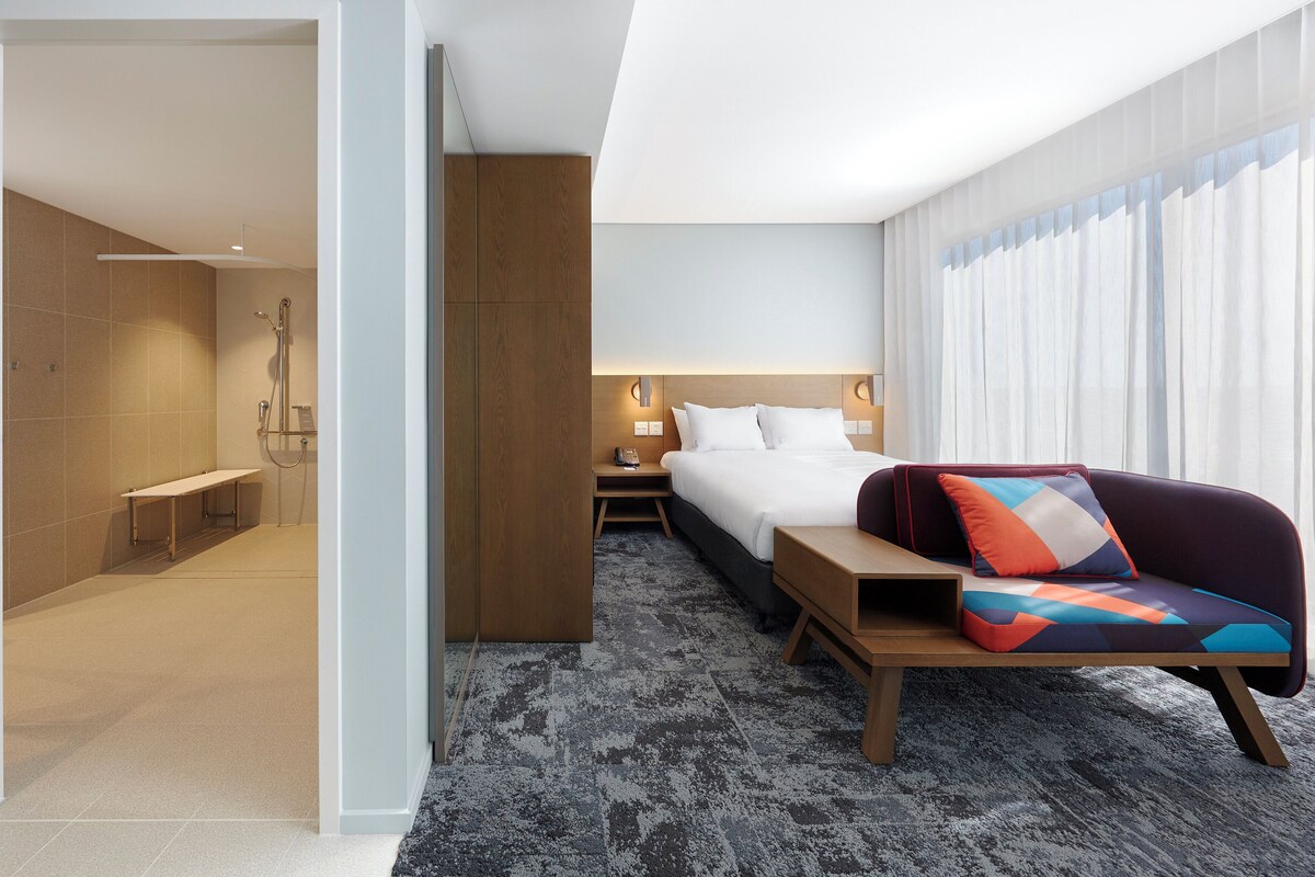 Hotelkamer met een bed, nachtkastje, douche en een groot raam met gordijnen.