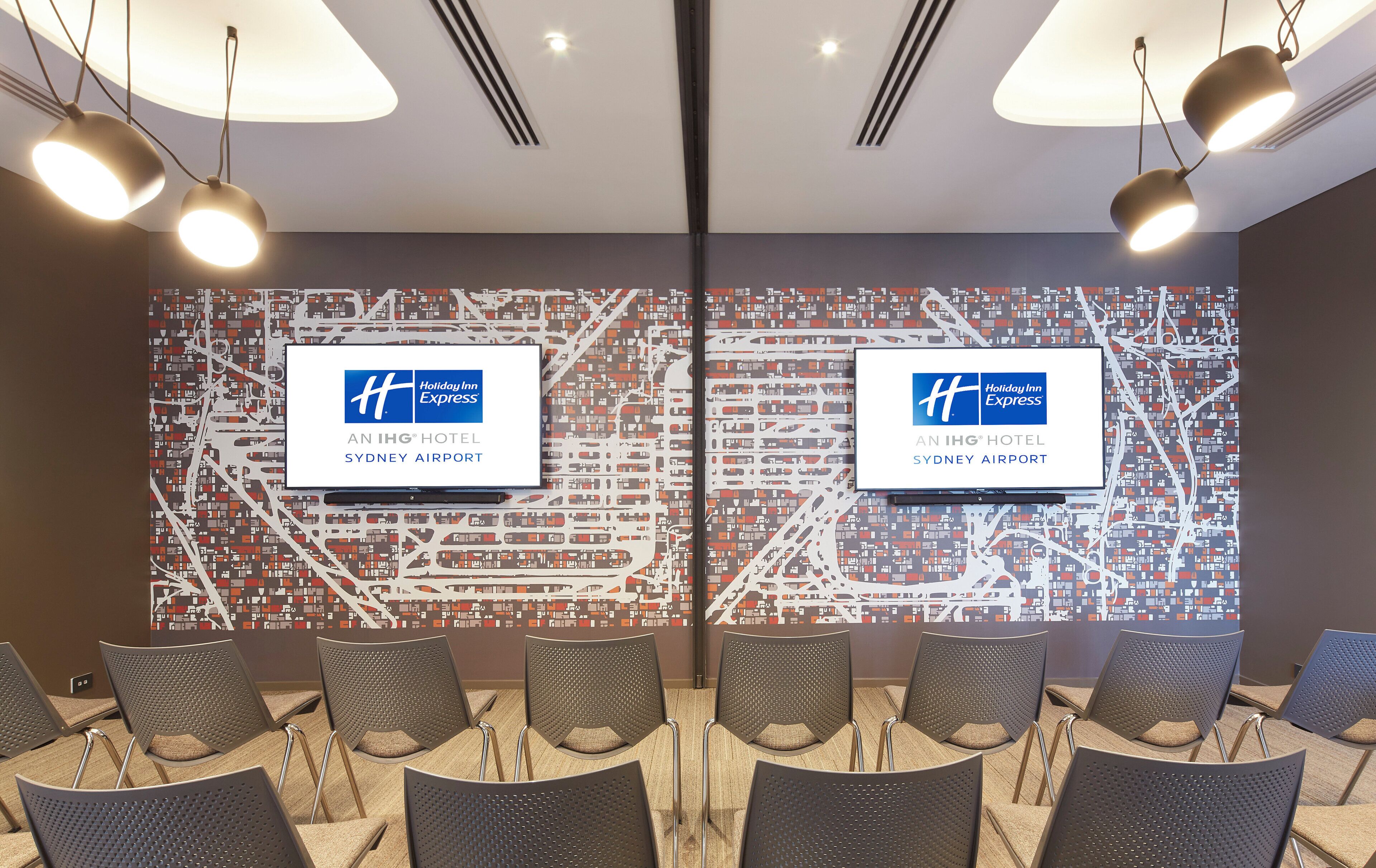 A conference room with two screens displaying the Holiday Inn Express logo, a map of Sydney Airport, and a row of chairs.