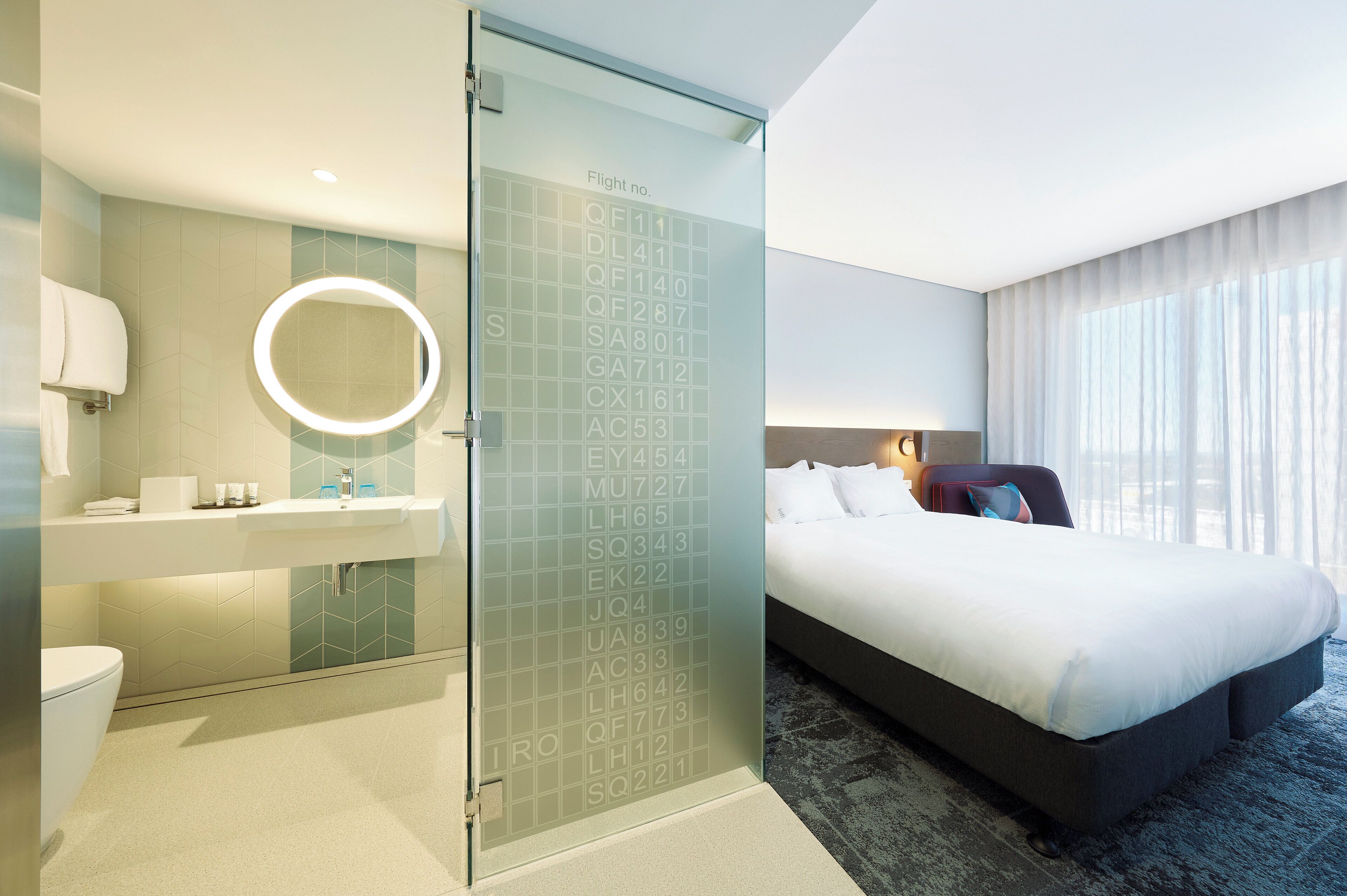 Habitación de hotel con cama, baño con espejo y puerta de vidrio con números de vuelo.