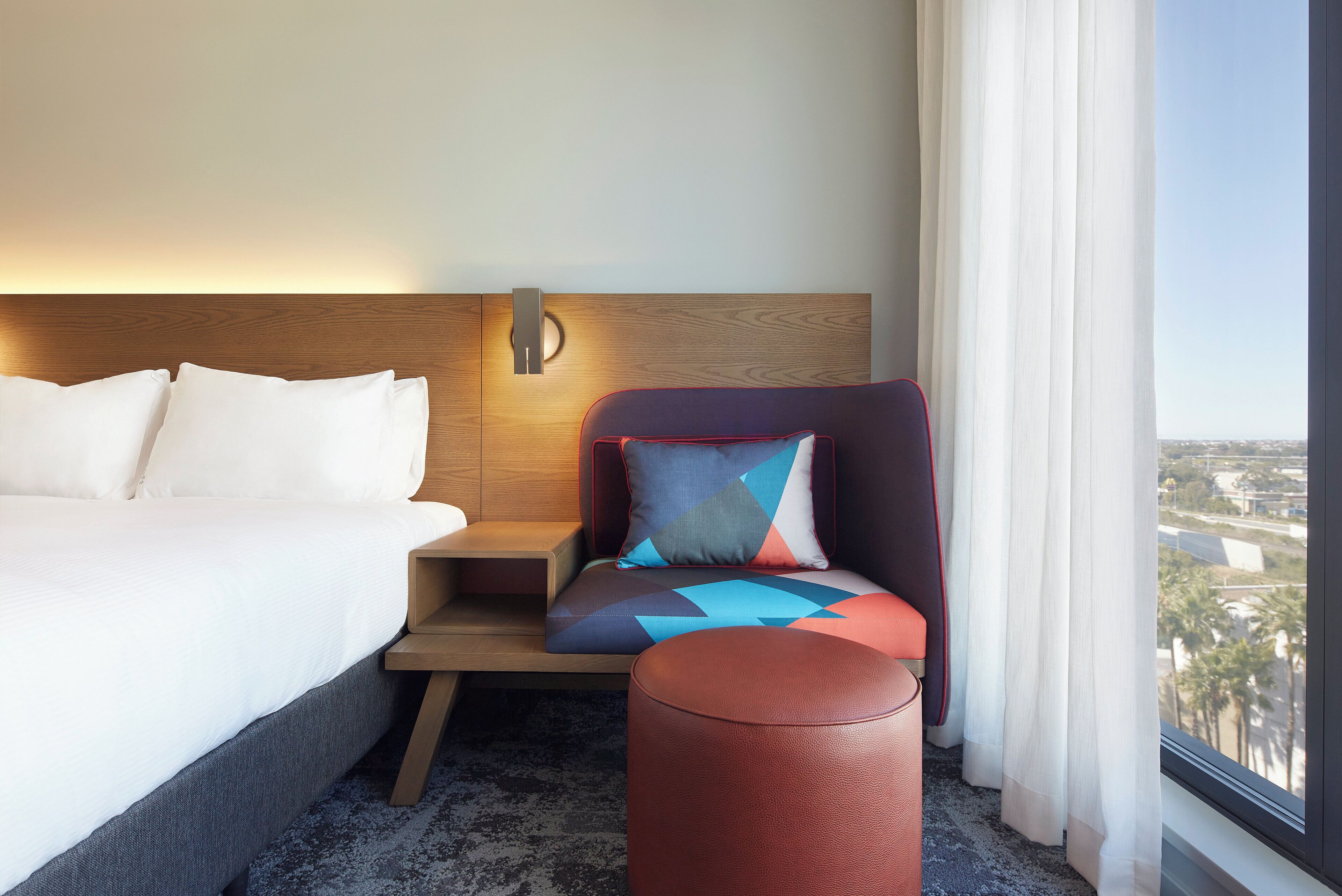 A hotel room with a bed, a nightstand, a chair with a geometric-patterned cushion, a round ottoman, and a view through a window.