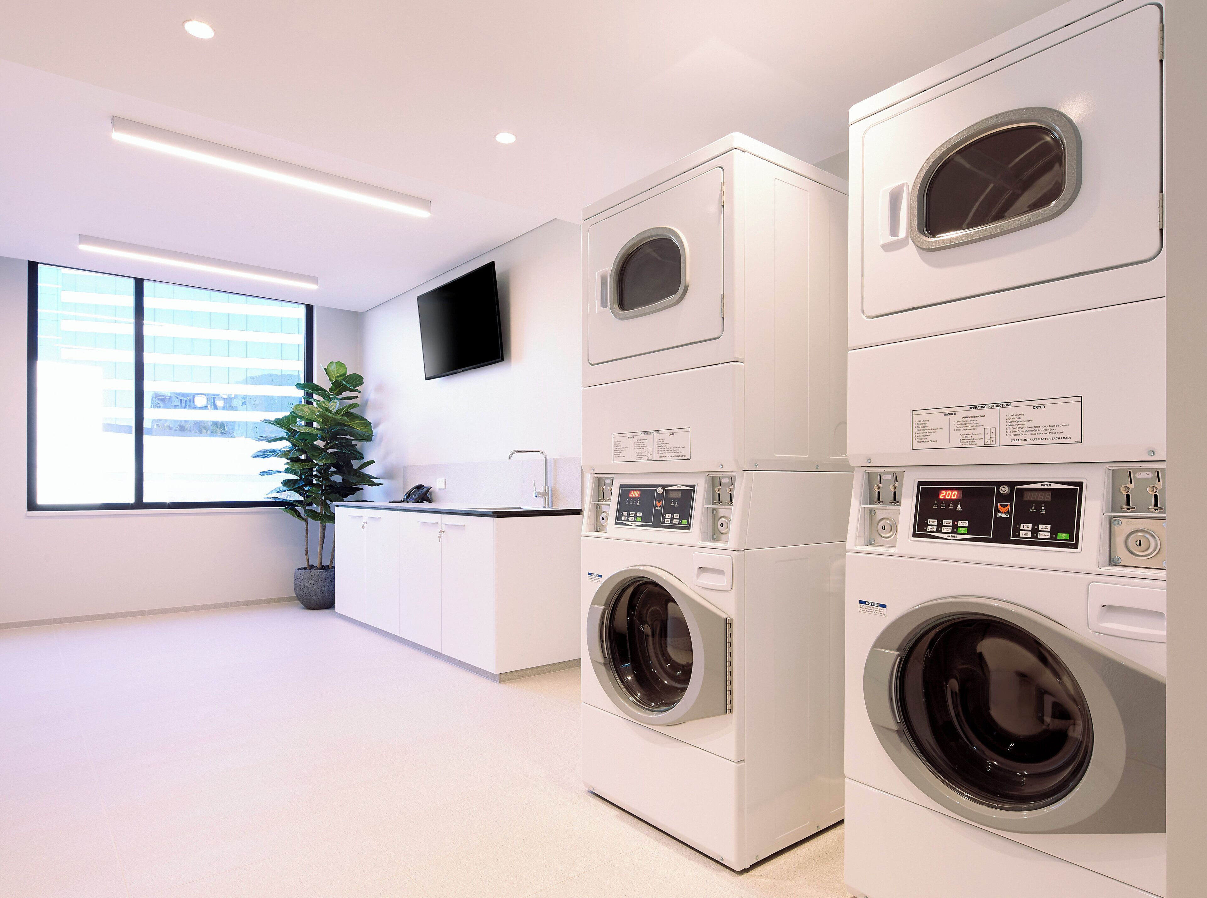 Une buanderie moderne équipée de machines à laver et de sèche-linge superposés, d’un évier, d’une plante en pot et d’un téléviseur à écran plat fixé au mur.
