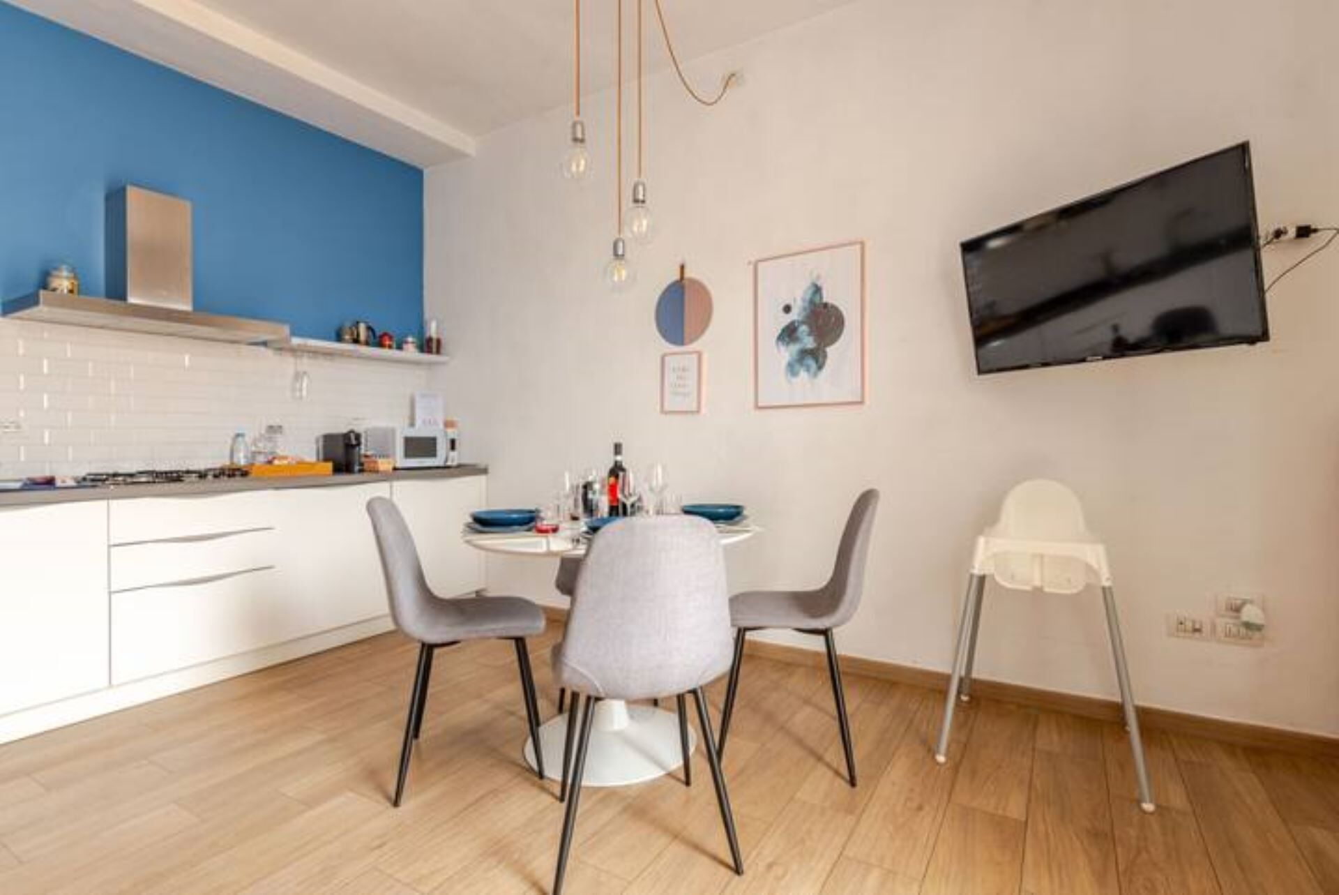 A modern kitchen with white cabinets, a blue accent wall, a dining table with four chairs, and a TV mounted on the wall.