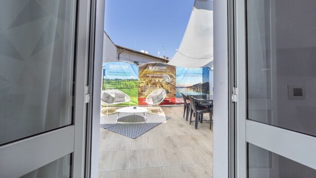A view through a glass door into a patio area with a white chair, a small table, and a banner advertising local attractions. - Boutique Apartment Eleganza (Chiusi Scalo)