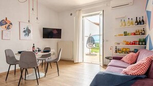 A modern living room with a dining area, a TV mounted on the wall, and a view of the outdoors through an open door. - Boutique Apartment Eleganza (Chiusi Scalo)