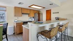 Cocina compacta con armarios de madera, refrigerador de acero inoxidable, microondas y una pequeña isla con taburetes de bar.