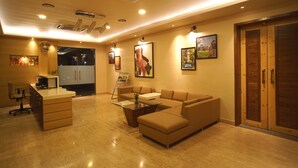 A modern living room with a sofa, coffee table, and a desk with drawers. There are ceiling lights and a door in the background. - Hotel Shree Kanha Residency (Prayagraj)