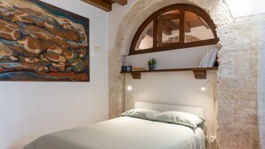 Une chambre à coucher avec un lit, un tableau, une étagère remplie de livres et une plante, et un mur en pierre.