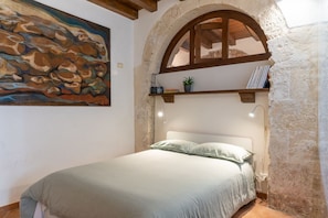 A bedroom with a bed, a painting, a shelf with books and a plant, and a stone wall.