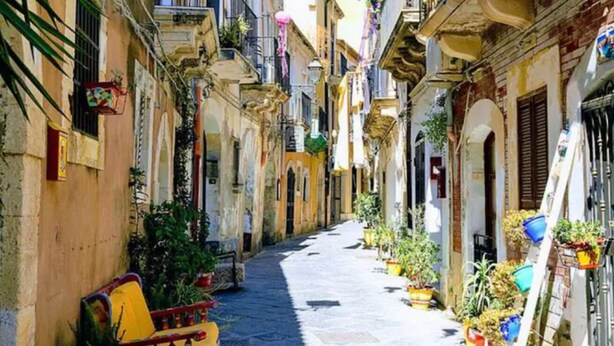 Une rue étroite, bordée de bâtiments des deux côtés, avec des plantes en pot et un banc.