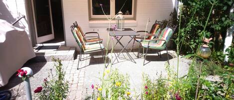 Un espace patio aménagé avec deux chaises à armature métallique et une petite table, entouré d’une variété de plantes et de fleurs en pot.
