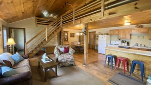 Ein gemütliches Wohnzimmer mit einer Couch, einem Couchtisch und einem gemusterten Sessel. Eine Treppe führt zu einer oberen Ebene. Der Küchenbereich umfasst Holzausstattung, einen Herd und Barhocker.