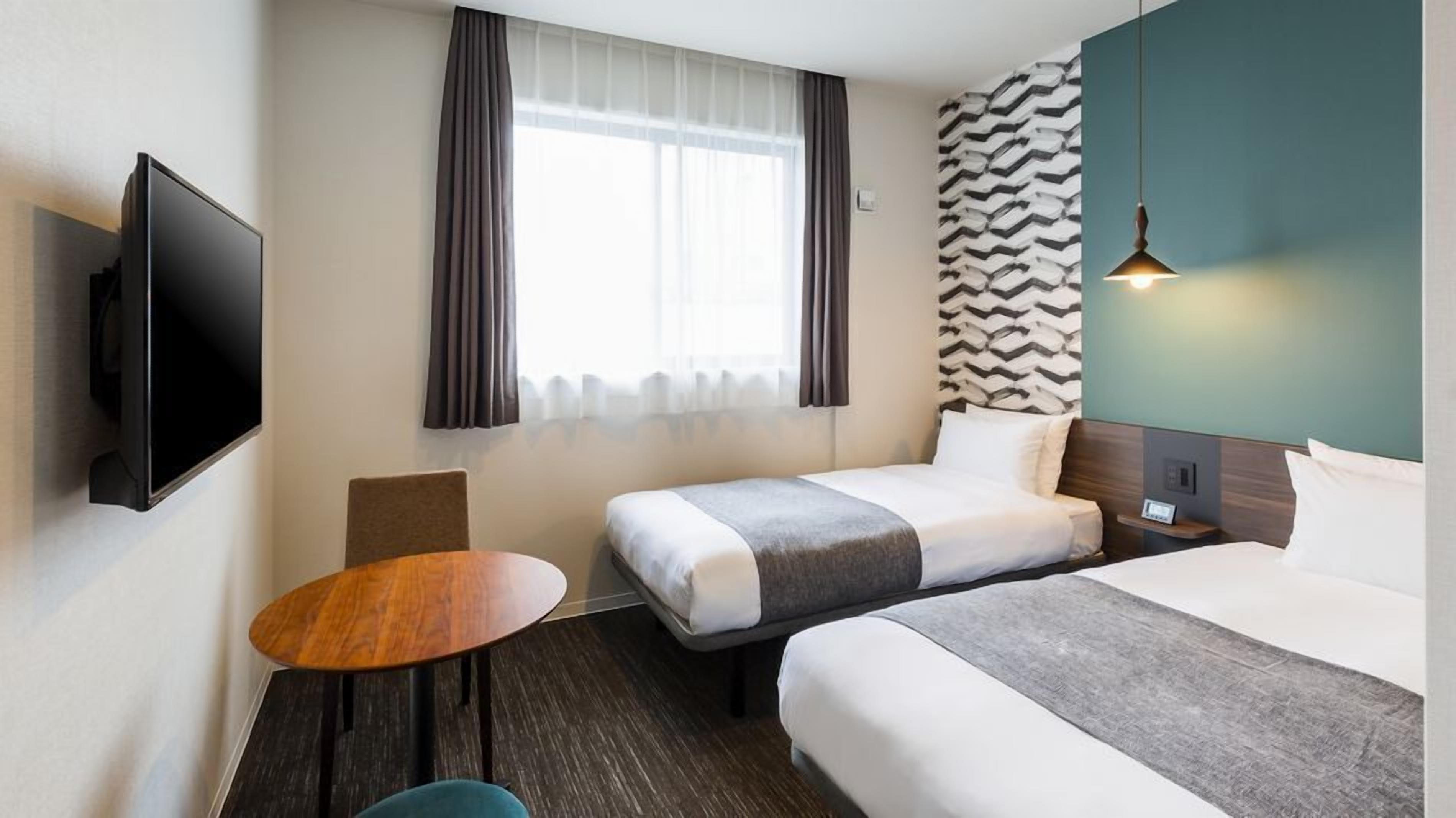A hotel room with two beds, a TV mounted on the wall, a small table, and a window with curtains.