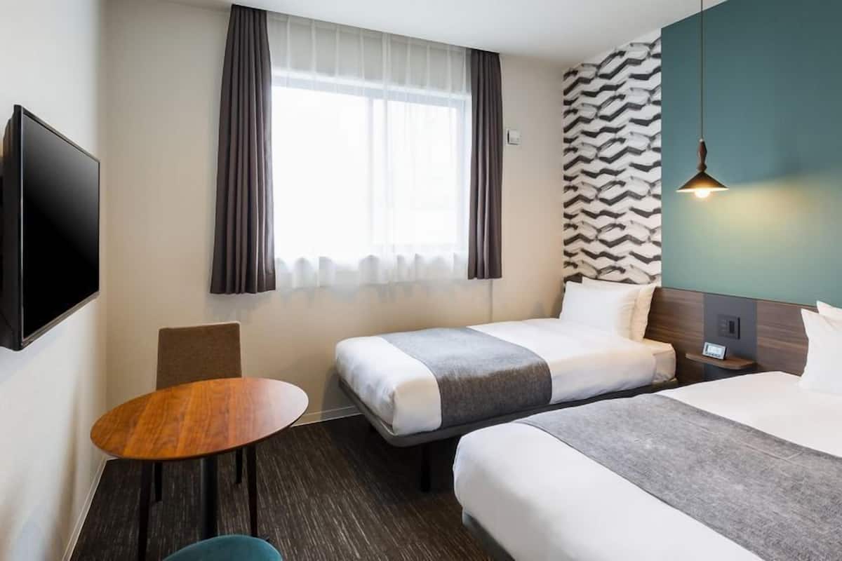 A hotel room with two beds, a TV mounted on the wall, a small table, and a window with curtains.