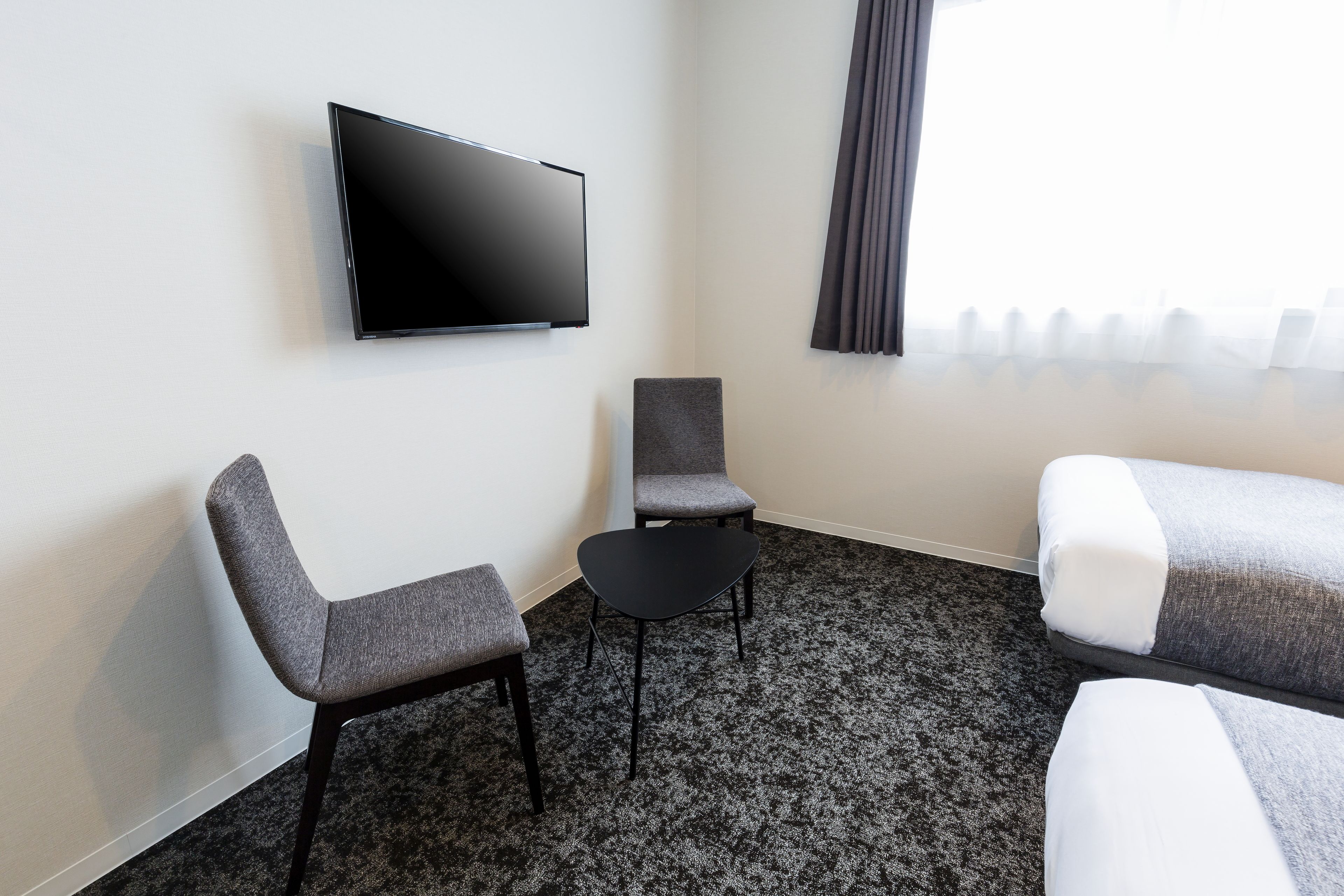 Une chambre d’hôtel équipée d’un téléviseur à écran plat, de deux fauteuils gris, d’une petite table noire et d’un lit avec une literie grise.