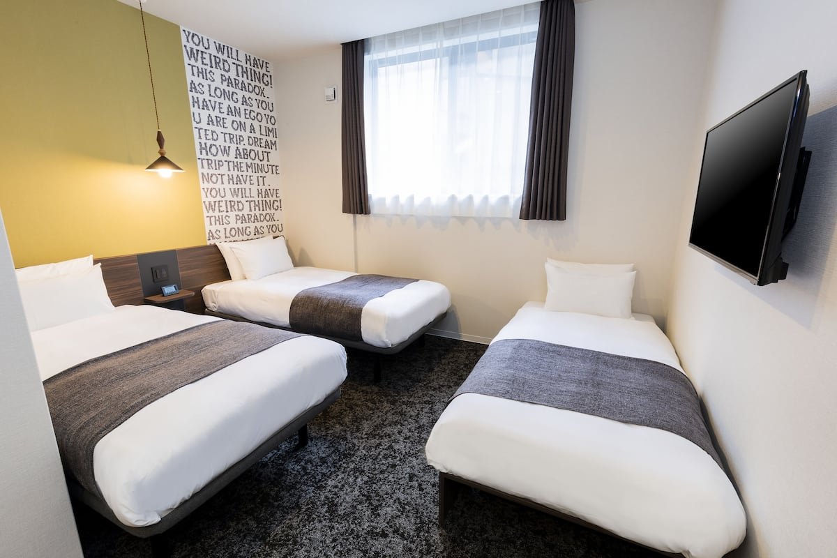 A hotel room with two single beds, a flat-screen TV, a wall-mounted lamp, and a large window with curtains.