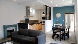 Una sala de estar moderna con un sofá gris, una mesa de comedor con sillas, una cocina al fondo y una pared azul decorativa con un reloj.