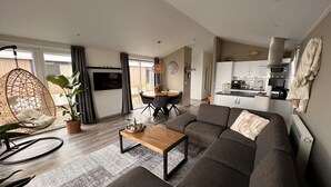 En moderne stue med sofa, hængegyngestol, spisebord og et køkkenområde med skabe og hårde hvidevarer.