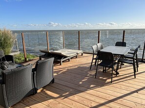 Eine Holzterrasse mit Gartenmöbeln, darunter ein Tisch und Stühle, mit Blick auf ein Gewässer.