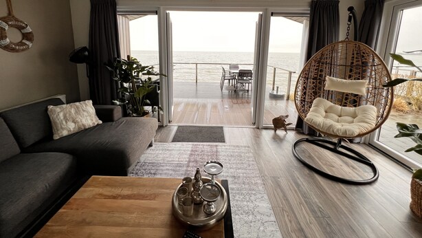 En moderne stue med sofa, sofabord og hængestol. Et stort vindue giver udsigt til havet.