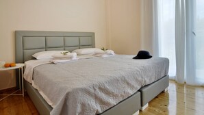 Una cama bien hecha con cabecera gris, una mesita de noche con lámpara y una ventana con cortinas traslúcidas.