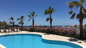 A swimming pool with a curved edge, surrounded by palm trees and pink flowers, with a clear view of the ocean in the background. - Stunning ocean view, Prime location, Exclusive design (Lagos)