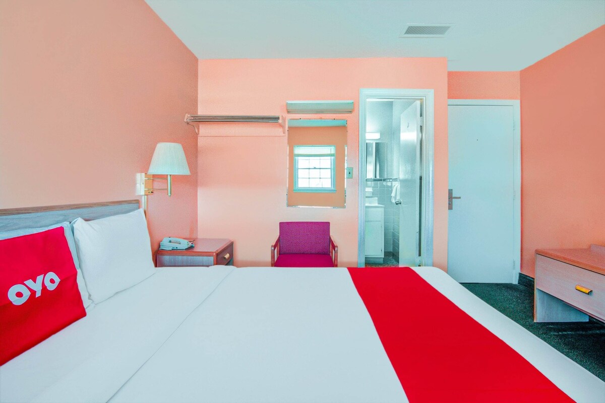 A hotel room with a bed, a nightstand, a chair, a desk, a lamp, and a door leading to a bathroom.