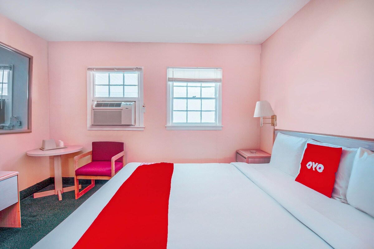 A hotel room with a bed, a chair, a small table, and two windows.