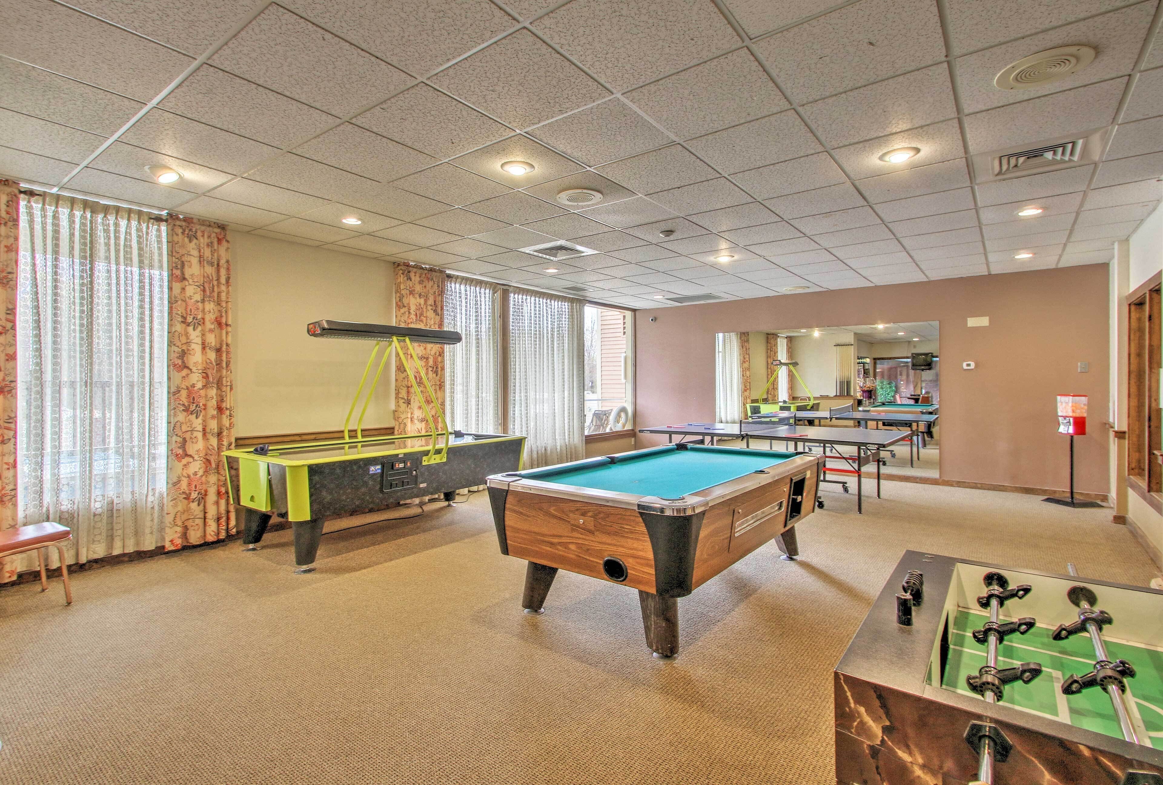 A pool table, foosball table, and a billiards table in a room with large windows and a carpeted floor.