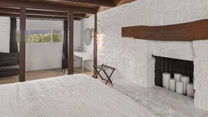 Une chambre avec un mur en briques blanches, un plafond en bois, une cheminée avec des bougies et une vue sur l’extérieur.