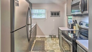 A compact kitchen with stainless steel appliances, a window with blinds, a pet feeding station, and a tiled floor. - Blowing Rock Home: 1 Block to Downtown! (Blowing Rock)
