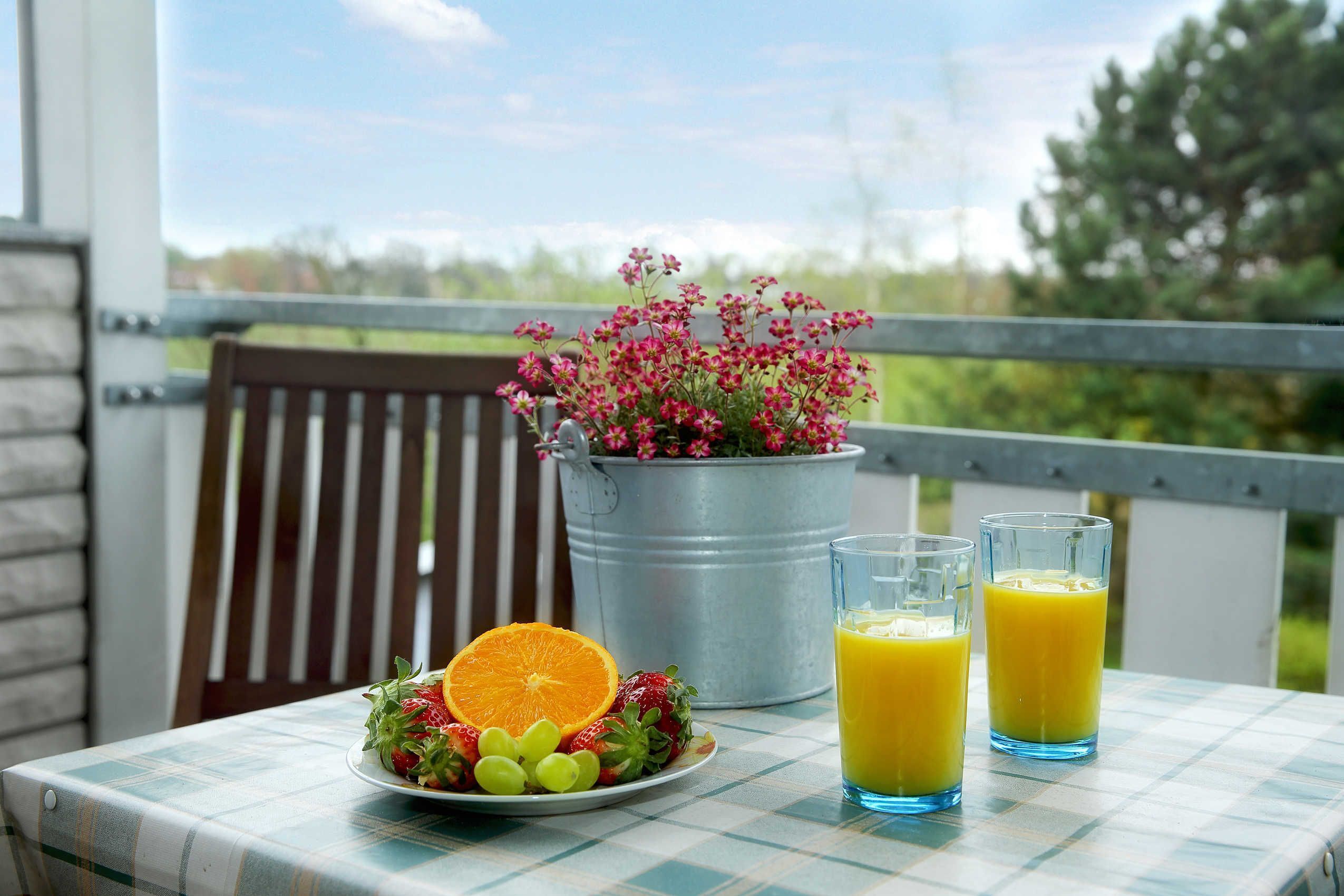 A table with a checkered tablecloth, a metal bucket with pink flowers, a plate of fruit, and two glasses of orange juice.