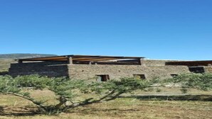 Un bâtiment rustique, doté d’un toit plat et d’une façade en pierre, entouré d’une végétation aride et surplombé d’un ciel dégagé.