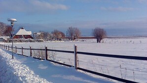 Un paysage enneigé avec une clôture, un bâtiment dont le toit est recouvert de neige et des arbres sans feuilles.