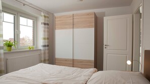 Un dormitorio con cama, ventana con cortinas, radiador, armario y puerta.