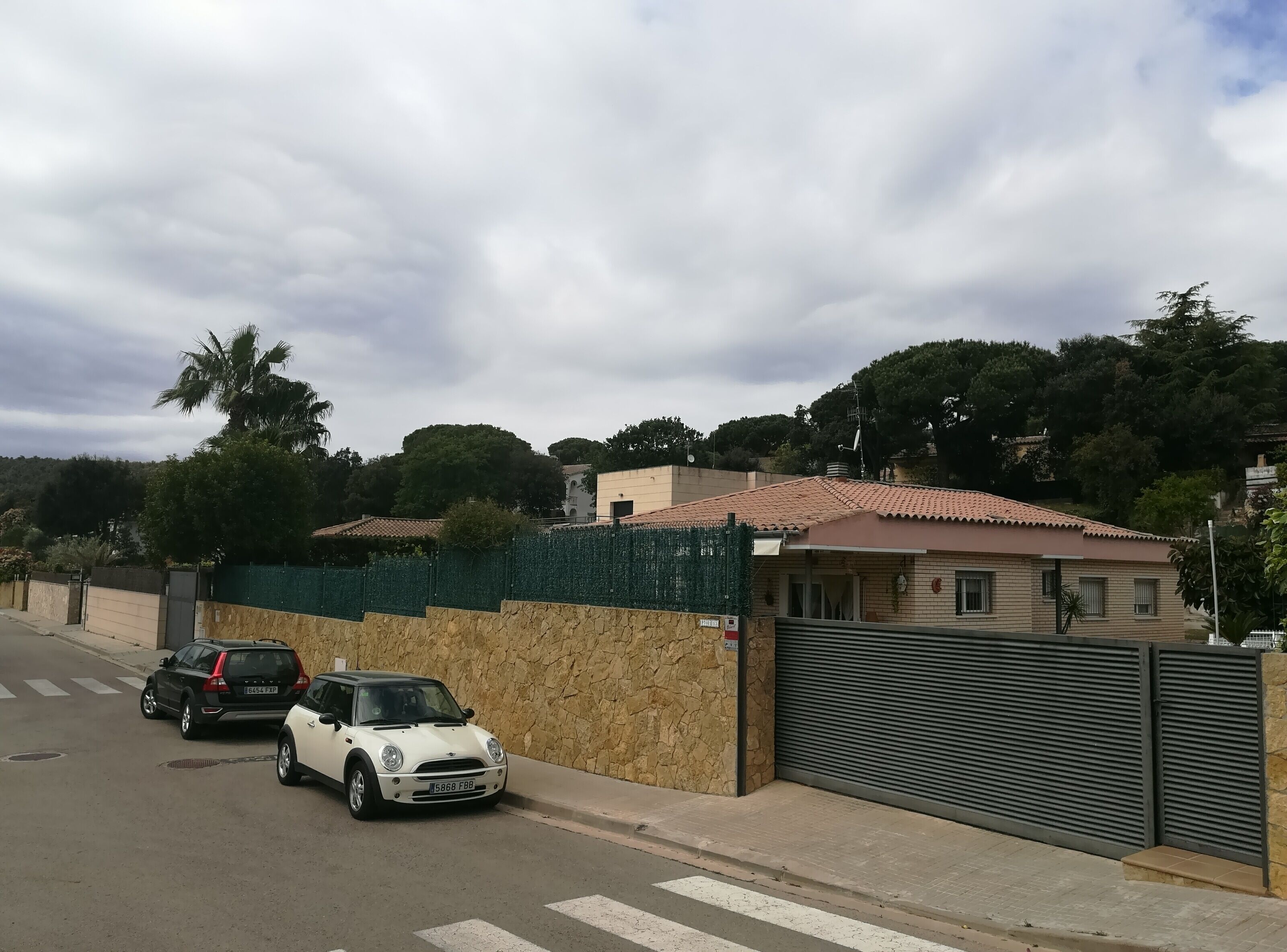 Eine Straße mit zwei parkenden Autos am Straßenrand, eine Steinmauer und ein Gebäude mit heruntergelassenem Rollo.