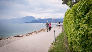 Ein gepflasterter Weg entlang eines Gewässers, im Hintergrund Berge.