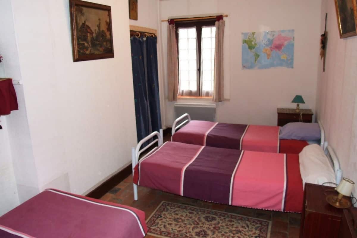 A bedroom with two beds, a window with curtains, a map on the wall, and a small table with a lamp.