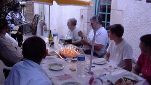 Um grupo de pessoas sentadas à mesa ao ar livre, com um bolo, garrafas de água e pratos.