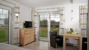 Une chambre d’hôtel moderne équipée d’un lit, d’une commode en bois, d’une télévision, d’une table avec une chaise, et offrant une vue sur l’extérieur grâce à de grandes portes coulissantes.