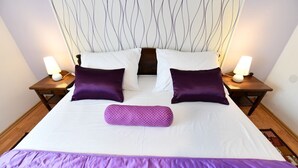Una cama bien hecha con ropa de cama blanca y morada, dos mesitas de noche con lámparas y un cabecero con diseño ondulado.