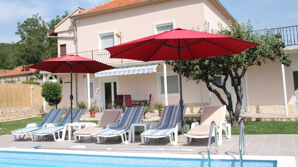 Un área junto a la piscina con sillones de descanso y sombrillas rojas, un edificio con tejado de tejas, y cielo despejado.