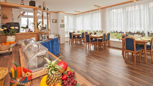Un comedor con mesas y sillas de madera, un frutero en la mesa y vistas al exterior a través de amplias ventanas.