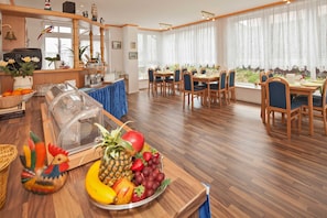 A dining area with wooden tables and chairs, a fruit bowl on the table, and a view of the outdoors through large windows. - Room 4 - Pension and Apartments Haus Antje (Heringsdorf)