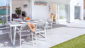 A modern outdoor dining area with a white table set for four, surrounded by glass doors leading to a patio. - Almendro by Interhome (Sta Cristina d'Aro)
