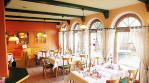A restaurant with multiple tables and chairs, large windows with curtains, and a warm color scheme.