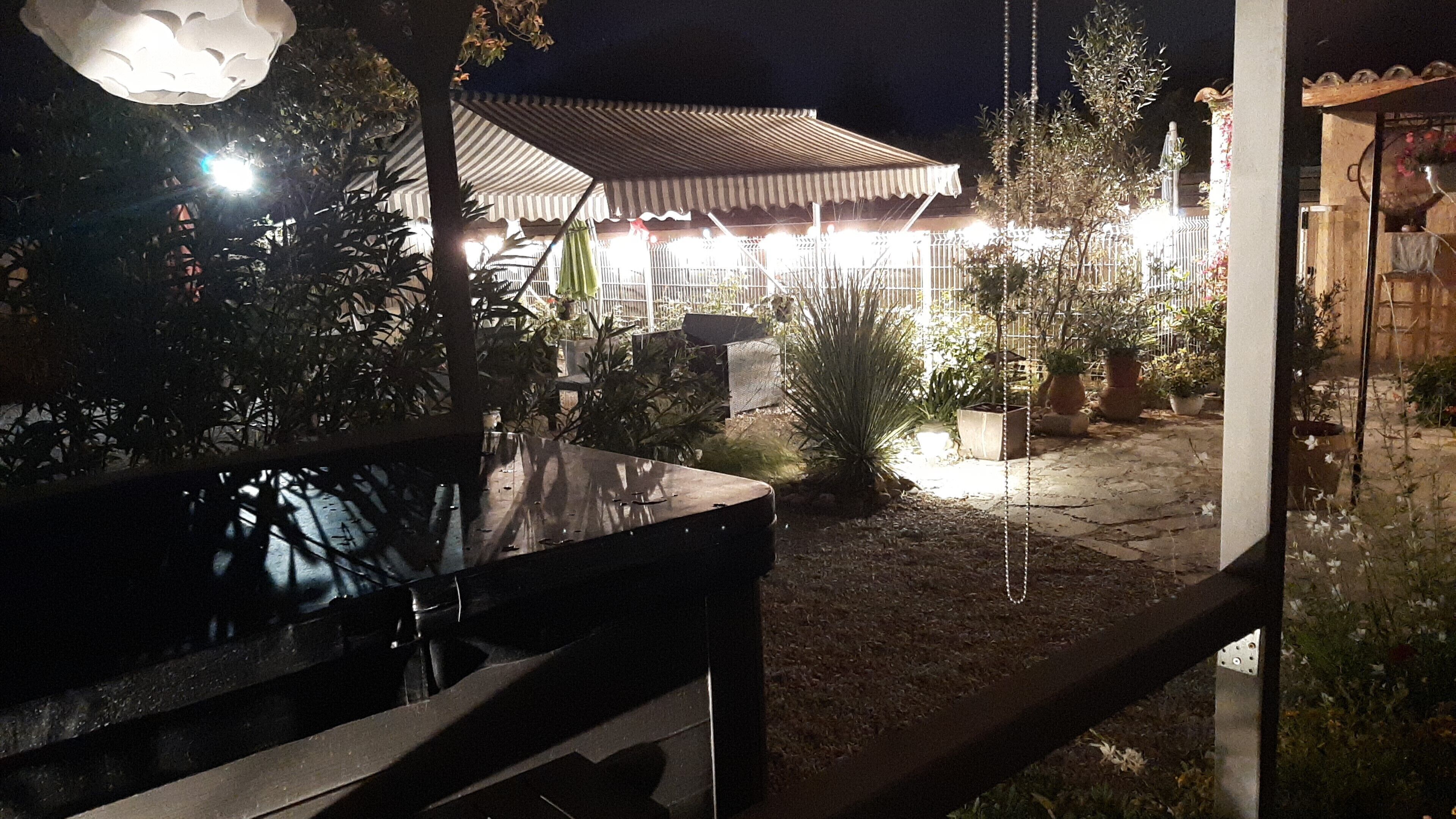 An outdoor patio area with a wooden bench, potted plants, and a building with a corrugated metal roof in the background.